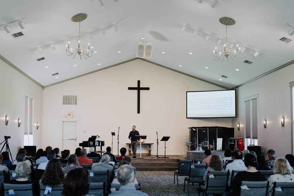 Comfortable modern church interior at Anderson Hills Church Salem Campus with congregation listening to a speaker at the pulpit, with a large cross and scripture projected behind.